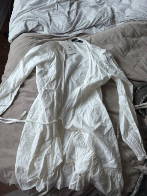 Vit omlottklänning med volang - Säljer en vit omlottklänning med långa ärmar och volangdetaljer. Klänningen har knytband i midjan och är tillverkad i ett lätt och luftigt tyg, perfekt för varmare dagar. Enkel och stilren design som passar till många tillfällen.