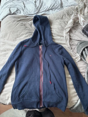 Mörkblå hoodie från Tommy Hilfiger - Snygg mörkblå hoodie från Tommy Hilfiger med dragkedja framtill och klassiska röda, vita och blå detaljer längs dragkedjan. Har huva med dragsko och en stor magficka. Liten logga på bröstet. Perfekt för en chill och stilren look.