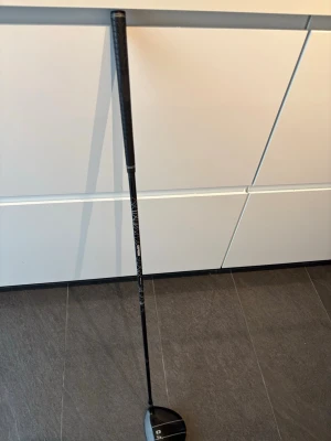 Svart open  driver  (Vänster) - En svart open  driver med silverfärgade linjer på klubbhuvudet. Skaftet är också svart med vita detaljer. Perfekt för dig som vill slå långa utslag på golfbanan.