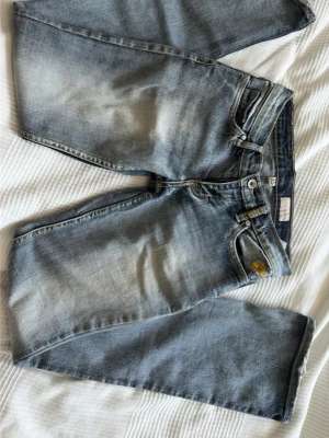 Ljusblå raka jeans med slitningar - Säljer ett par ljusblå jeans med raka/utsvängda ben och snygga slitningar. Jeansen har klassisk femficksmodell, gylf med dragkedja och knapp samt detaljer i gult vid ena fickan. Perfekta för en avslappnad och trendig look.