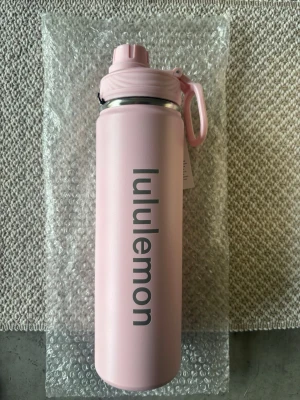 Rosa vattenflaska från Lululemon - Snygg rosa vattenflaska från Lululemon med praktiskt lock och handtag. Flaskan är tillverkad i rostfritt stål och har en matt finish med Lululemon-loggan tryckt i grått längs sidan. Perfekt att ha med till gymmet eller skolan.