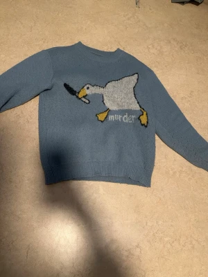 Blå stickad tröja med gås-motiv - Blå stickad tröja med ett stort motiv av en gås som håller i en kniv och texten 'murder' på framsidan. Tröjan har rund hals, långa ärmar och ribbstickade muddar. Perfekt för dig som gillar unika och roliga plagg.