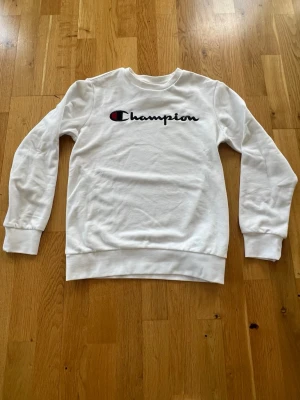 Vit sweatshirt från Champion  - 150/155 cl .Säljer en clean vit sweatshirt från Champion med klassisk logga broderad i svart, rött och blått på bröstet. Tröjan har rund hals, långa ärmar och ribbade muddar vid ärmslut och nederkant. Perfekt för en avslappnad och sportig stil.Nyskick, bara provad