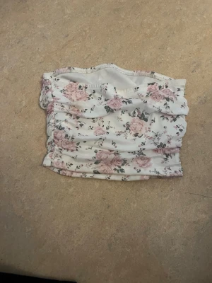 Vit blommig bandeau topp - Säljer en söt bandeau topp i vitt med rosa blommönster och små gröna blad. Toppen har rynkade detaljer på sidorna och är axelbandslös, perfekt för sommaren. Materialet känns mjukt och stretchigt.
