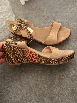 Beige kilklackade sandaler  - Snygga sandaler med beige mockaimitation och färglatt kilklack . Remmar med spänne runt ankeln ger en cool och bekväm look. Perfekta för dig som vill ha något unikt och trendigt till sommaren. Helta nya, aldrig använda men har ingen prislapp!