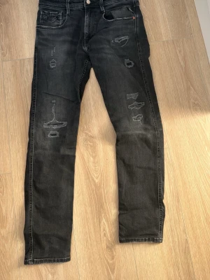 Svarta Replay jeans med slitningar - Säljer ett par svarta jeans från Replay med coola slitningar och distressed detaljer på benen. Jeansen har fem fickor, smal passform och klassisk knappgylf. Storlek w31 o l30 