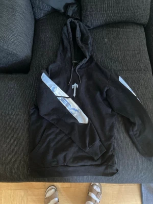 Trapstar hoodie  - Säljer en svart hoodie med stor huva och dragsko. På ärmarna finns blå och vita grafiska tryck och på bröstet en pil-liknande detalj. Baksidan har ett stort tryck i blått och vitt med tredelad design. Hoodien har känguruficka och ribbade muddar.