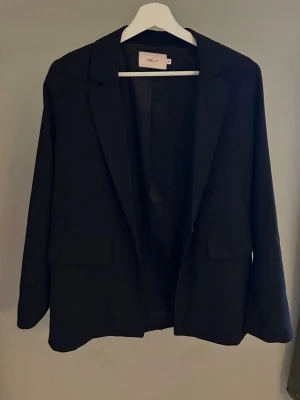 Svart kavaj från Nelly - Stilren svart kavaj från Nelly med klassisk krage och två fickor framtill. Rak passform och helfodrad insida. Perfekt för en trendig och clean look.