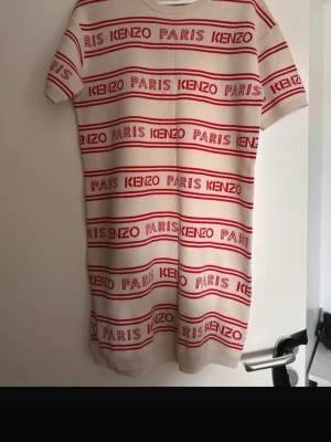 Randig kortklänning från Kenzo - Säljer en beige och röd kortklänning från Kenzo med randigt mönster och logotypen 'KENZO PARIS' tryckt över hela klänningen. Klänningen har korta ärmar och är tillverkad i mjuk bomull. Perfekt för dig som gillar streetstyle och vill sticka ut.