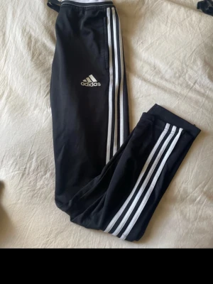 Svarta Adidas träningsbyxor - Säljer ett par svarta träningsbyxor från Adidas med de klassiska vita ränderna längs benen och Adidas-logga på låret. Byxorna har resår i midjan och är tillverkade i ett mjukt, stretchigt material som passar perfekt för sport och träning. Dom har slitage mellan bena men perfekt för den som kan sy🪡🧵