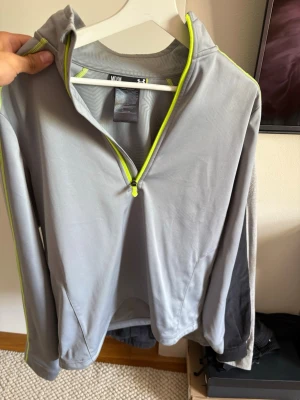Golf vind tröja från under armour - Snygg grå träningshoodie från Under Armour med halv dragkedja framtill och limegröna detaljer längs dragkedjan och ärmarna. Tillverkad i ett lätt och stretchigt material som passar perfekt för löpning eller gym. Huva och fickor på sidorna.