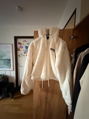 Vit teddyjacka med huva - Mysig vit teddyjacka med dragkedja framtill och huva. Jackan har dragsko i nederkant och en liten patch på bröstet. Perfekt för kalla dagar när du vill vara både varm och stilren. Materialet är fluffigt och mjukt för maximal komfort.