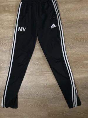 Svarta Adidas träningsbyxor - Säljer ett par svarta träningsbyxor från Adidas med vita ränder längs sidorna och Adidas-logga på ena benet. Byxorna har resår i midjan och smal passform. Perfekta för träning eller chill. Initialerna MV är tryckta på ena benet.