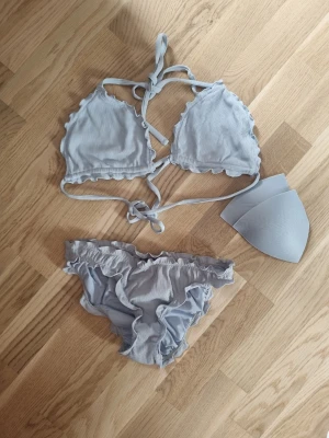 Ljusblå bikini med halterneck strl S/M - Supersöt ljusblå bikini med volangdetaljer och ribbad struktur. Överdelen har triangelformade kupor med knyt i nacken och ryggen. Bikinitrosan har matchande volangkant och scrunch i bak.. Uttagbara inlägg medföljer. Överdelen är i strl M och underdelen är i S. Är använda ca 7 ggr. Nypris för båda är ca 330.