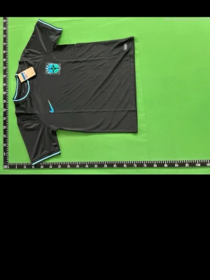 Svart Brasilien fotbollströja med blå detaljer  - Snygg svart fotbollströja från nike med ljusblå logga och matchande blå detaljer vid krage och ärmslut. Tröjan har ett tryckt klubbmärke på bröstet och är tillverkad i ett lätt och ventilerande material som passar perfekt till träning.