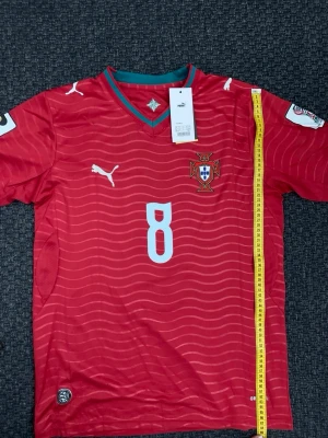 Bruno Fernandes nummer 8 Röd Portugal fotbollströja Puma - Snygg röd Portugal fotbollströja från Puma med nummer 8 på bröstet och landslagets emblem. Tröjan har vita detaljer, korta ärmar och V-ringad krage med grönt och rött. Perfekt för match eller träning.
