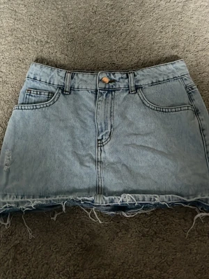 Ljusblå jeanskjol med fransig kant - Säljer en ljusblå jeanskjol med rå, fransig kant nertill och klassiska fem fickor. Kjolen har bälteshällor och knappstängning framtill. Perfekt för dig som gillar en avslappnad och trendig stil.