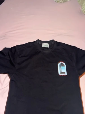 Svart t-shirt från Casablanca - Säljer en svart t-shirt från Casablanca med ett tryck på bröstet i blått och rött. Skön passform och klassisk rund halsringning. Perfekt för dig som gillar stilrena plagg med en twist.