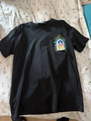 Svart t-shirt med tryck från Casablanca - Snygg svart t-shirt från Casablanca med färgglatt tryck på bröstet där det står 'Avenida Casablanca' och ett motiv av en byggnad omgiven av gröna blad. Klassisk rund hals och korta ärmar. Perfekt för en chill och stilren look.