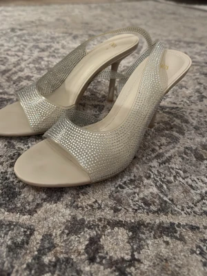 Glittriga slingback-pumps från ZARA - Säljer ett par snygga pumps från ZARA i storlek 39. Skorna är beige med glittriga stenar över hela ovandelen och har öppen tå samt slingback-rem. Klacken är smal och elegant, och materialet känns som syntet med en lyxig finish.