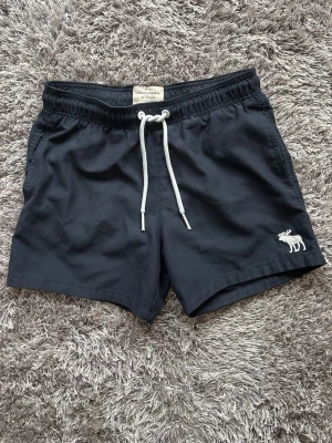 Badbyxor från Abercrombie & Fitch - Mörkblå badbyxor från populära märket Abercrombie & Fitch med vit snörning och en broderad älglogga på vänster ben. Klassisk passform med elastisk midja och bakficka med patch. Perfekta för stranden eller poolen.    