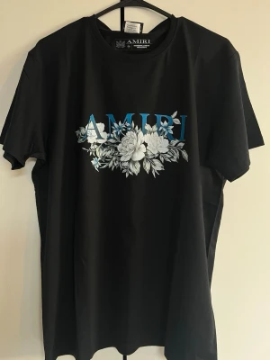 Svart AMIRI t-shirt med blommor - Snygg svart t-shirt från AMIRI med stort blått och vitt blommönster och AMIRI-tryck på bröstet. Klassisk rund hals och korta ärmar. Perfekt för dig som vill ha en stilren men ändå unik look. Tillverkad i mjuk bomull för skön känsla.