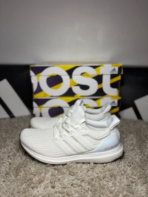 Adidas Ultra Boost vita sneakers - Snygga Adidas Ultra Boost sneakers i helvitt med stickad ovandel och klassiska tre ränder på sidan. Skorna har en bekväm Boost-sula och svart yttersula med vitt mönster. Perfekta för dig som gillar stilrena och sportiga skor.