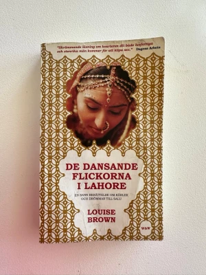 De dansande flickorna i Lahore - En fängslande bok av Louise Brown med omslag i vitt och guld, prydd med ett foto av en kvinna i traditionell indisk smyckning. Boken handlar om kärlek och drömmar till salu i Lahore. Perfekt för dig som gillar gripande och verklighetsbaserade berättelser.