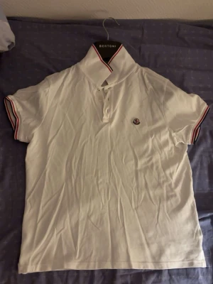 Vit moncler pikétröja - Äkta vit Moncler pikétröja med korta ärmar och klassisk krage. Kragen och ärmsluten har rödblå ränder och det finns en liten broderad logga på bröstet. Tröjan har knäppning framtill och är gjord i ett mjukt material.