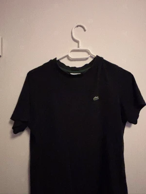 Svart t-shirt från Lacoste - Svart t-shirt från Lacoste med klassisk krokodillogga på bröstet. T-shirten har rund hals med grön kant och korta ärmar. Perfekt för en stilren och enkel look.