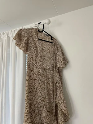 Beige mönstrad klänning från SHEIN - Säljer en beige kortklänning från SHEIN med brunt prickigt mönster. Klänningen har volangdetaljer, omlottmodell och korta, vida ärmar. Perfekt för sommarens alla tillfällen och superlätt att styla med accessoarer.