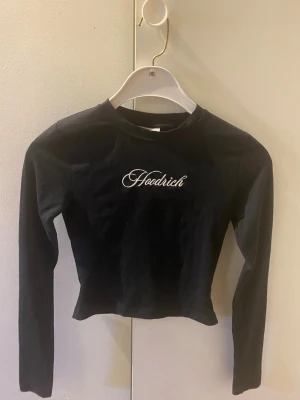 Svart långärmad topp från Hoodrich - Svart långärmad croppad topp från Hoodrich med vit broderad logga på bröstet. Tight passform och rund halsringning. Perfekt för dig som gillar streetwear och vill ha en enkel men snygg topp till din outfit.