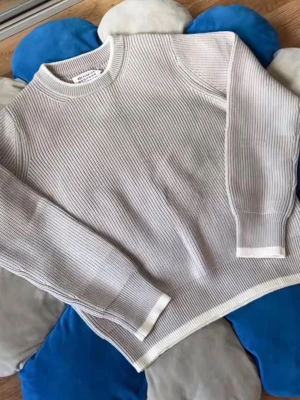 Ljusgrå stickad tröja Maison Margiela - Säljer en stilren ljusgrå stickad tröja från Maison Margiela. Tröjan har ribbstickad struktur, rund halsringning och vita kontrastkanter vid hals, ärmslut och nederkant. Perfekt för dig som gillar minimalistisk och modern design.