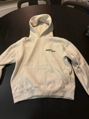 Grå Essentials Fear of God hoodie - Snygg grå hoodie från Essentials Fear of God med svart logga på bröstet och stor text på ryggen. Klassisk känguruficka framtill och stor huva. Tillverkad i mjukt bomullsmaterial som känns skönt mot huden. Perfekt för en avslappnad streetwear-look.