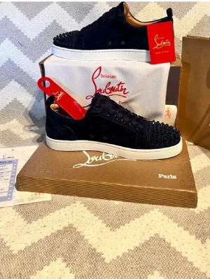 Christian Louboutin svarta sneakers - Svarta sneakers från Christian Louboutin med klassiska röda sulor och svarta nitar på tån. Skorna har en låg profil, svart mocka och vita gummisulor. Snygg och edgy design som sticker ut med exklusiva detaljer och ikonisk look.