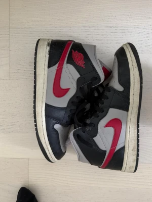 Air Jordan 1 Mid svart/röd/grå - Säljer ett par Air Jordan 1 Mid sneakers i svart, grått och rött. Skorna har klassisk röd swoosh, svart snörning och den ikoniska Air Jordan-loggan på sidan. Tillverkade i läder med bekväm vadderad insida. Perfekta för dig som gillar streetwear och snygg design.