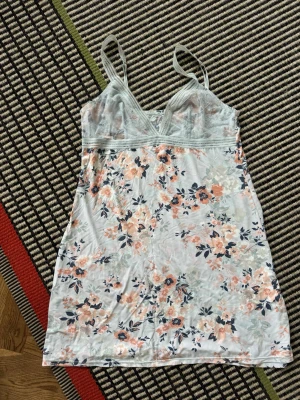 Blommig slip-dress med spetsdetaljer - Slip-dress med ljusblå bas och blommigt mönster i persika, blått och vitt. Spets upptill och justerbara axelband. Snygg själv eller till jeans.