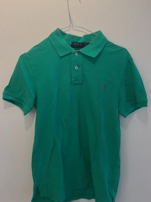 Grön pikétröja från Polo Ralph Lauren - Barn storlek, 160. Klassisk grön pikétröja från Polo Ralph Lauren med korta ärmar och krage. Tröjan har två knappar framtill och den ikoniska lilla loggan broderad på bröstet i rosa. Tillverkad i bomull för en bekväm känsla.