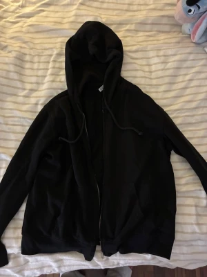 Svart zip-hoodie med dragsko - En klassisk svart hoodie med dragkedja framtill och huva med dragsko. Hoodien har långa ärmar, ribbade muddar och två fickor framtill. Perfekt för en avslappnad och stilren look.