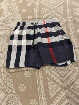 Rutiga badbyxor med dragsko - Snygga badbyxor med klassiskt rutigt mönster i svart, vitt, grått och en röd rand. De har elastisk midja med svart dragsko och sidofickor samt en bakficka. Materialet är lätt och snabbtorkande, perfekt för stranden eller poolen.