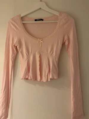 Ljusrosa topp med spets från Gina Tricot - Supersöt ljusrosa topp från Gina Tricot med långa ärmar, spetsdetaljer längs ringningen och framtill samt en liten rosett vid urringningen. Modellen är croppad och har en feminin, figurnära passform. Perfekt att styla med jeans eller kjol.