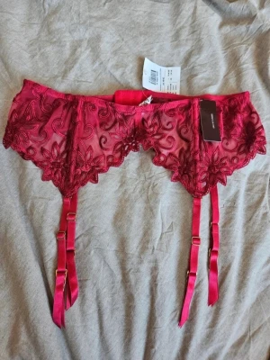 Röd spetsig strumpebandshållare - Säljer en snygg röd strumpebandshållare i transparent spets med blommigt broderi och justerbara band. Perfekt för att ge outfiten en extra touch. Materialet är tunt och lätt, med fina detaljer och dekorativa kanter.