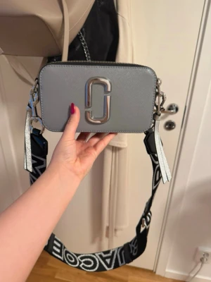 Grå Marc Jacobs Snapshot handväska - Snygg grå handväska från Marc Jacobs med silverfärgad logga framtill och bred, svart axelrem med vit text. Väskan har två dragkedjor upptill och detaljer i vitt och silver. Tillverkad i tåligt konstläder med stilren, boxig form.