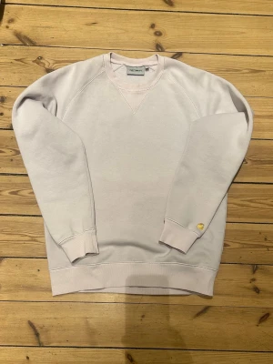 Ljuslila sweatshirt från Carhartt - En stilren ljuslila sweatshirt från Carhartt med rund halsringning och ribbade muddar vid ärmslut och nederkant. Tröjan har en liten gul Carhartt-logga på vänster ärm och är tillverkad i mjukt bomullsmaterial. 