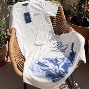 Vit t-shirt med blått tryck - Säljer en vit t-shirt med blått tryck och texten 'Mariel' på bröstet. Baksidan har ett stort konstnärligt motiv med natur, båt och vinflaska i blå toner. Klassisk passform och rund hals. Perfekt för dig som gillar unika prints.