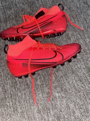 Röda Nike Mercurial fotbollsskor - Säljer ett par röda Nike Mercurial fotbollsskor med svart swoosh och högt skaft i strumpliknande material. Skorna har snörning och dobbar för bra grepp på planen. Snygg design med detaljer i svart och text på sidan. Perfekta för dig som vill sticka ut på fotbollsplanen.