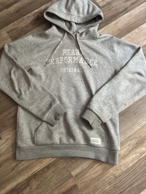Grå Peak Performance hoodie - Snygg grå hoodie från Peak Performance med broderad text på bröstet. Klassisk känguruficka framtill, ribbade muddar och dragsko i huvan. Perfekt för chill dagar och enkel att matcha med jeans eller joggers.