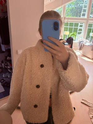 Krämvit teddyjacka med knappar - Mysig krämvit teddyjacka med tre stora svarta knappar framtill. Jackan har en boxy passform och är gjord i fluffigt teddy-material som ger en trendig look. Perfekt för att hålla värmen under kyliga dagar och passar till de flesta outfits.