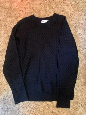 Svart stickad tröja från Kappahl - Säljer en svart stickad tröja från Kappahl i storlek XL. Tröjan har ett diskret diamantmönster, rund halsringning och ribbade muddar vid ärmslut och nederkant. Perfekt för dig som gillar enkel och stilren design.