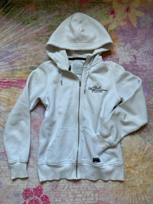Snygg Vit zip hoodie från Peak Performance - Säljer en enkel vit zip hoodie från Peak Performance med dragkedja framtill och broderad logga på bröstet!😍Hoodien har justerbar huva med snören, två fickor fram och ribbade muddar. Perfekt för en avslappnad men ändå snygg stil✨Den är skön på insidan och passar bra när du vill ha något lätt över din tröja men inte bli för varm🙏Använd men i bra skick utan fel. Passar bra för storlek XS! Hör av vid funderingar eller bud❤️